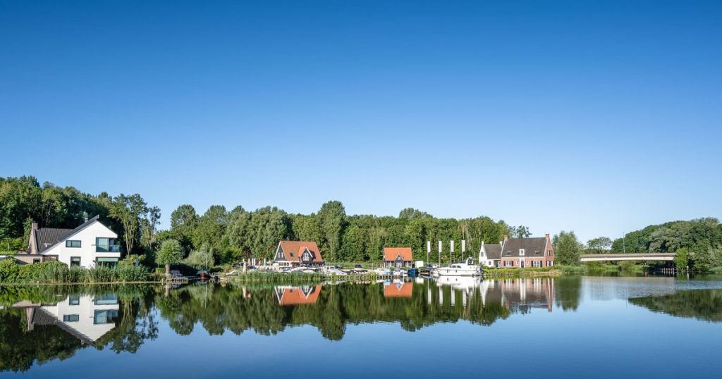 Sloep vaart over een stille plas bij Reeuwijk tussen rietkragen en houten vakantiehuisjes onder een blauwe lucht