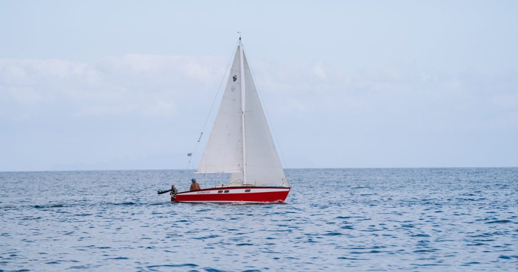 Een zeilboot met rode romp vaart over kalm water onder een blauwe lucht