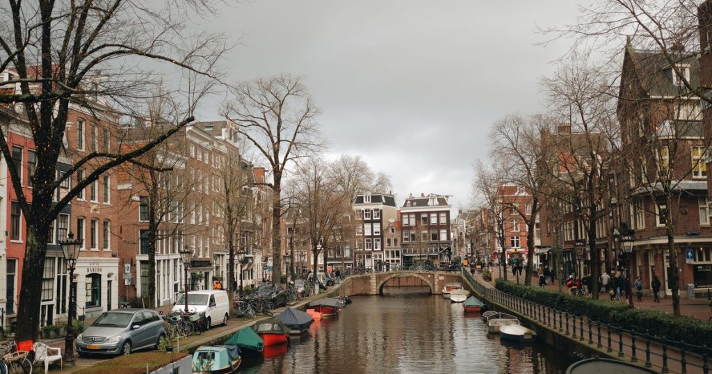 Uitzicht op een Amsterdamse gracht met boten, herenhuizen en een brug.