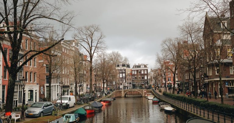 Uitzicht op een Amsterdamse gracht met boten, herenhuizen en een brug.