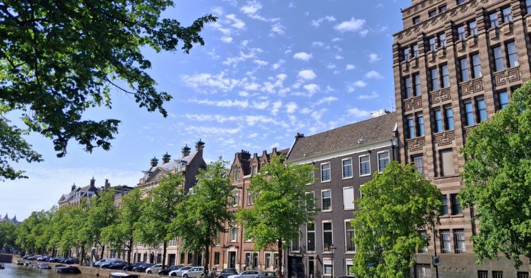 Een gracht met historische panden en bomen onder een heldere zomerlucht