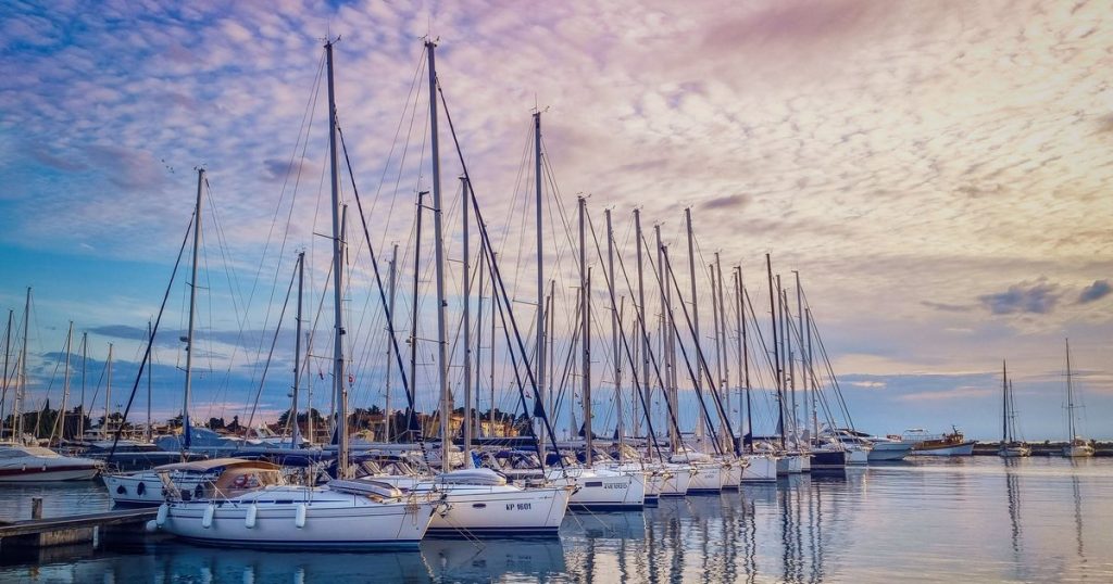Rustige jachthaven met afgemeerde zeilboten die weerspiegelen in het water bij zonsondergang, met een kleurrijke lucht.